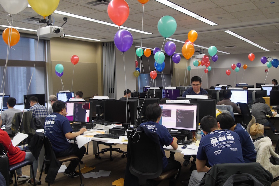 ICPC contestants working at computers in a contest room filled with colorful balloons.