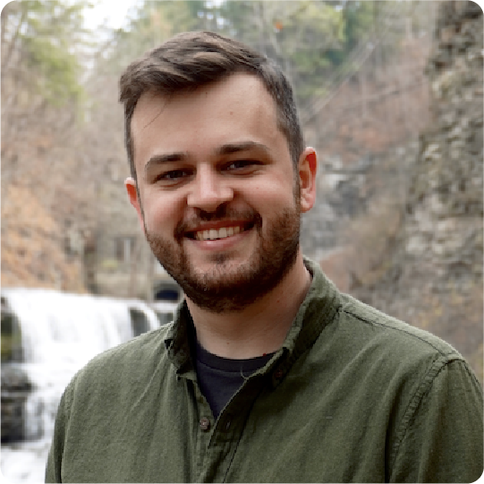 A color photo of a man smiling for a photo in front of a small waterfall.