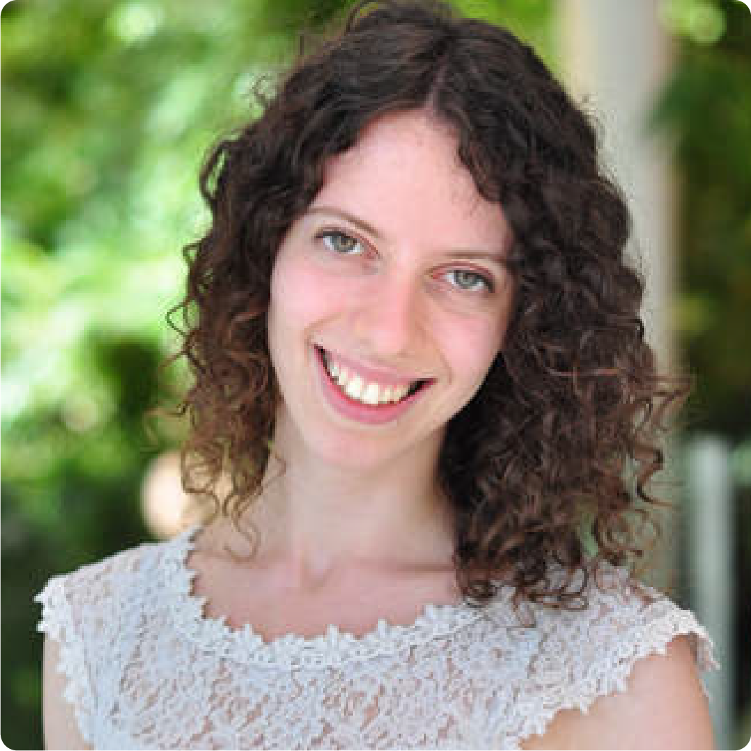 A color photo of a woman with curly hair, smiling for a photo outdoors.