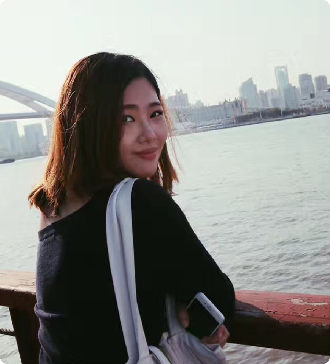 A color photo of a woman standing by a body of water, smiling for a photo.