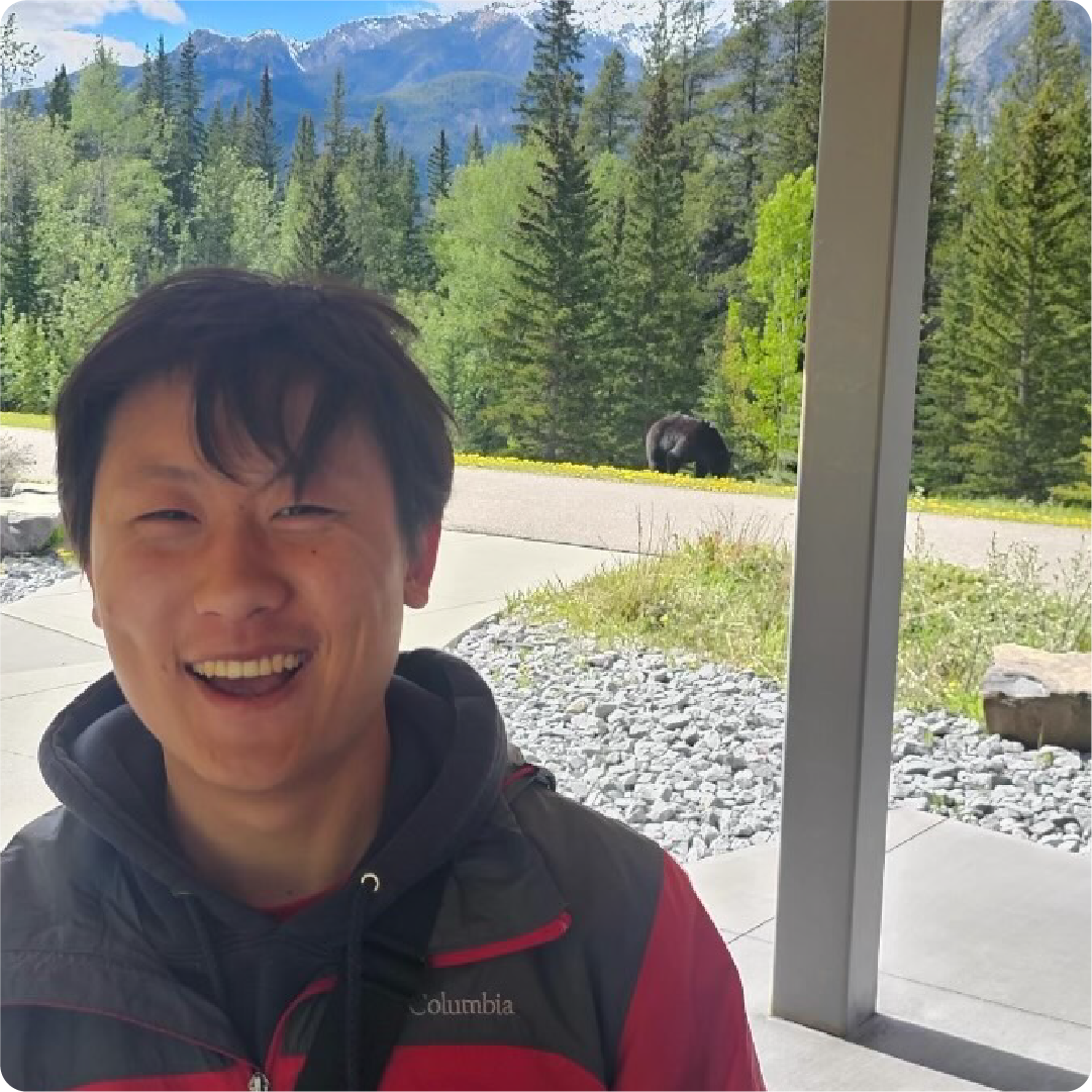 A color photo of a man standing near a forest with a bear visible in the background.