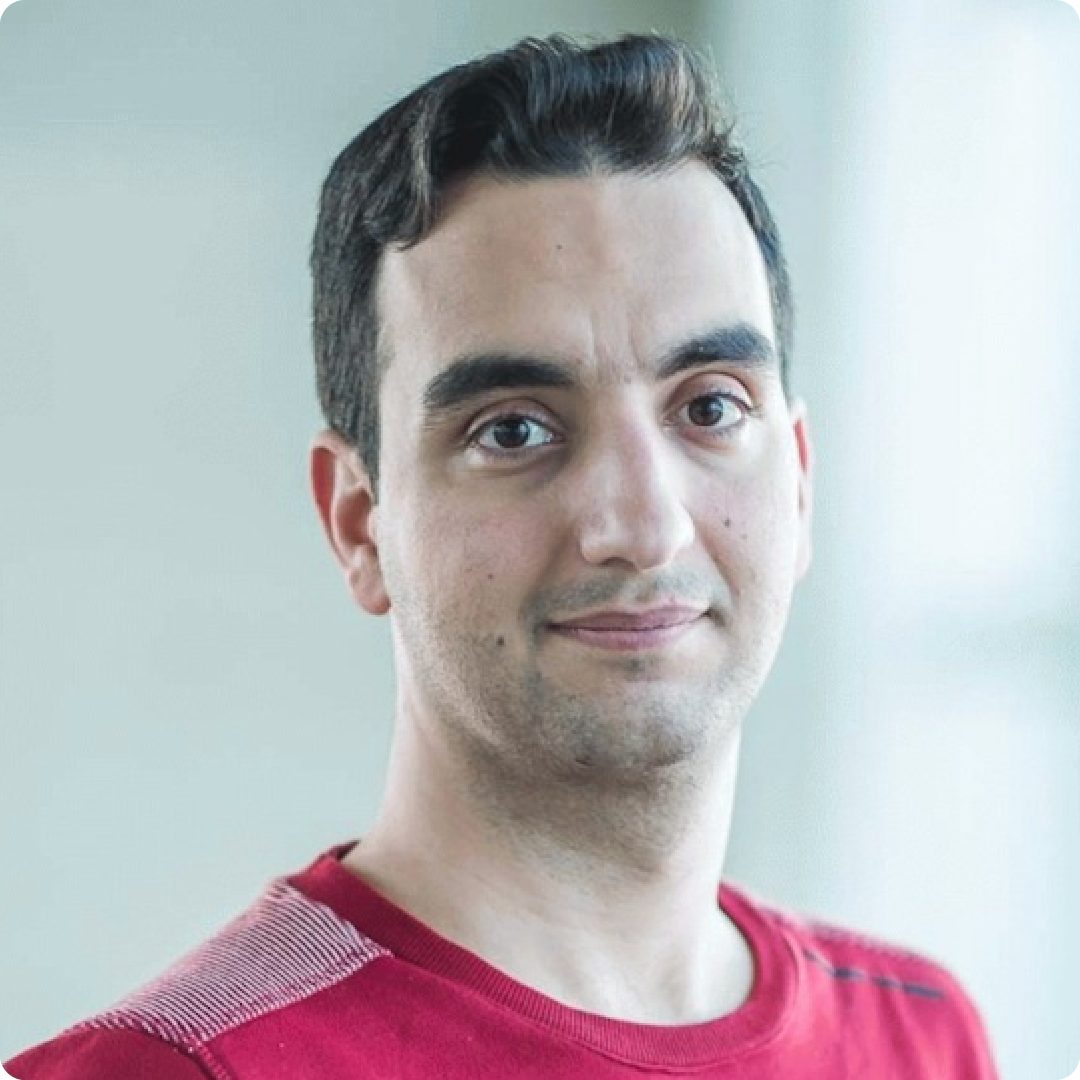 A color photo of a man smiling for a photo wearing a red t-shirt.