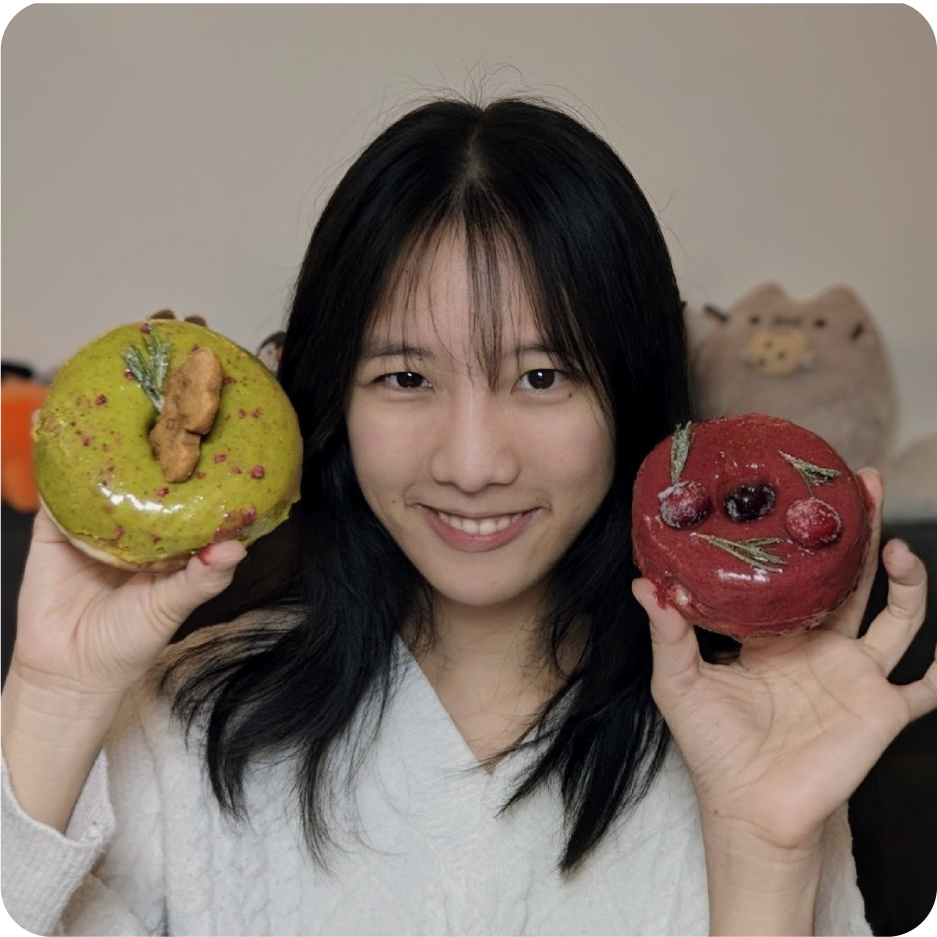 A color photo of a woman smiling for a photo while holding a donut in each hand.