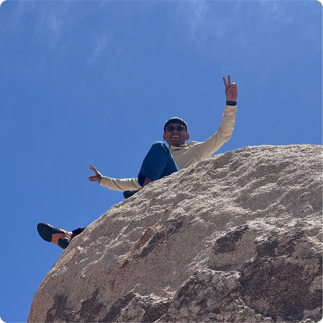 A color photo of a man sitting on top of a boulder.