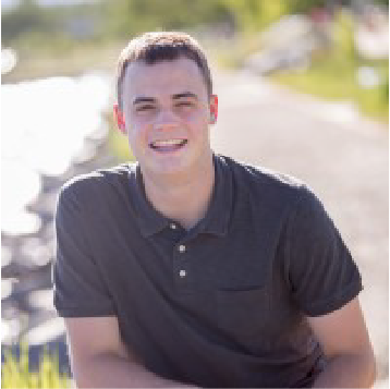 A color photo of a man smiling for a photo by a stream.