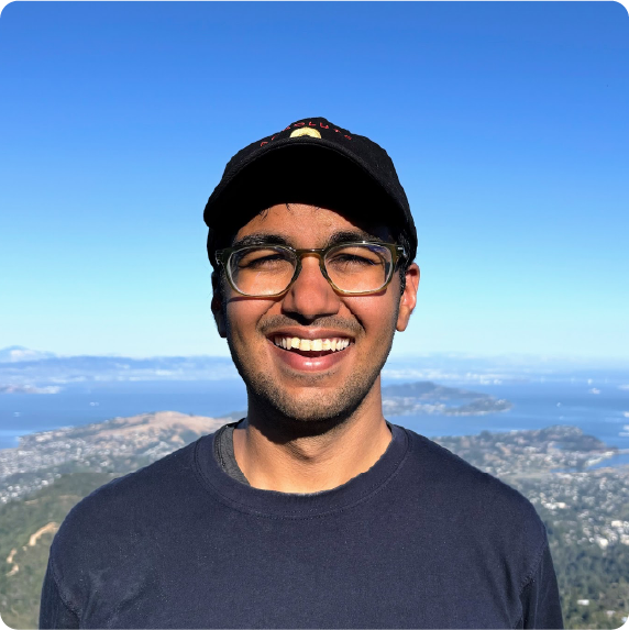 A color photo of a man smiling from a mountain top overlooking a bay.