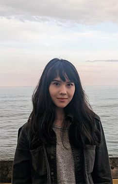 A color photo of a woman standing by a large body of water.