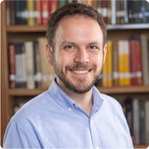 A color photo of a man smiling for a photo in front of a book case.