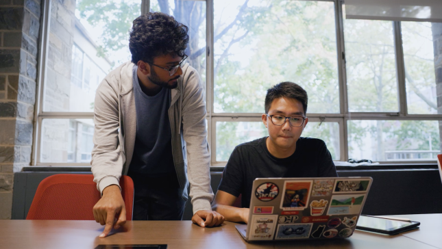 two students (one standing, one sitting) look at computer laptop