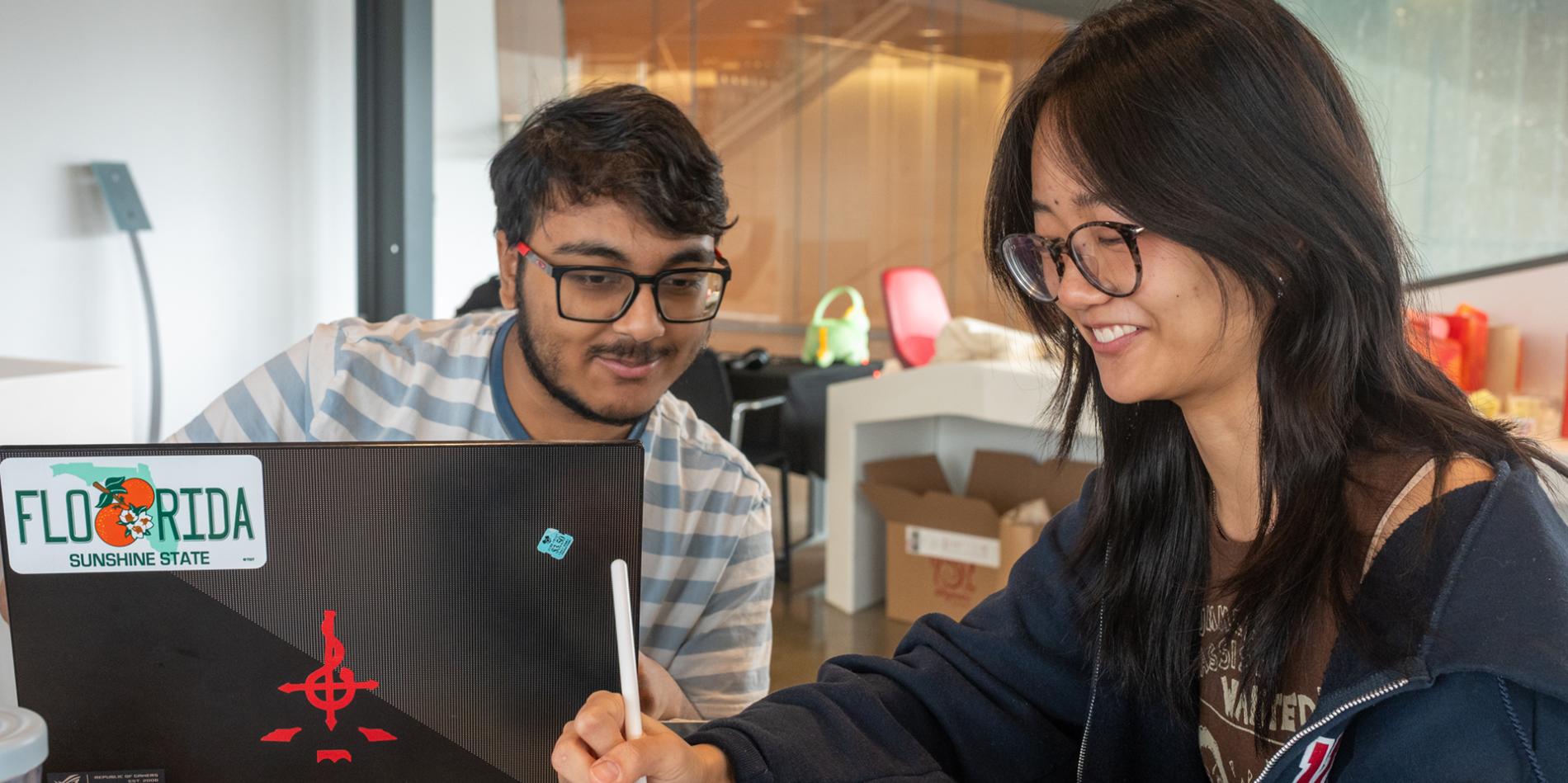 Two students sit around a laptop computer with a Flordia license plate bumper sticker. They wear glasses and smile, looking down at a tablet. 