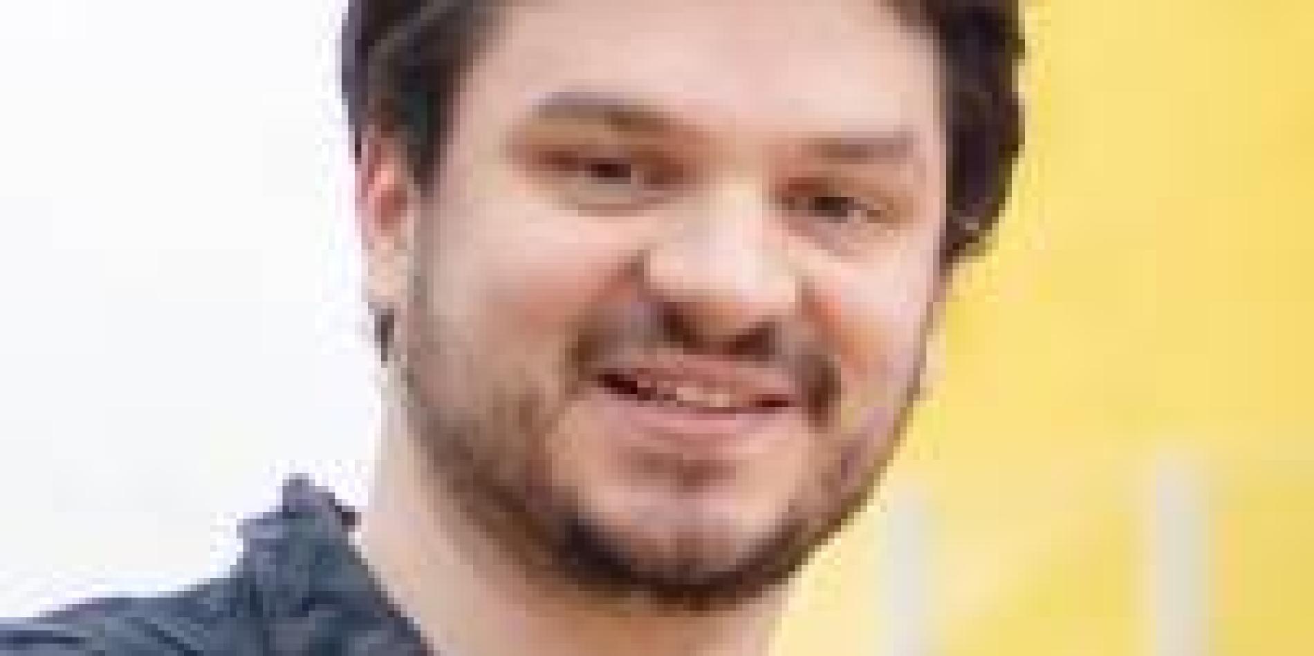 a man with short brown hair and a beard smiles in front of a white and yellow background