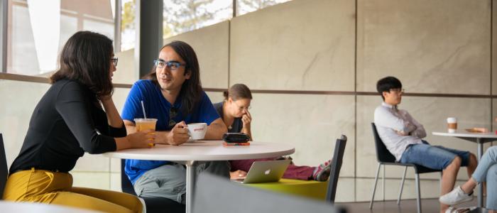 a male and female sit at a table with coffee outside with other people sitting at tables in the background