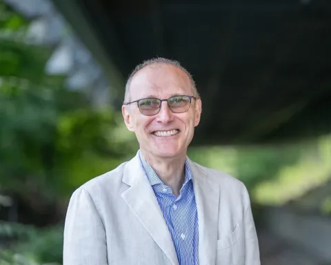 Man with glasses, short hair, blue shirt and tan jacket smiling at the camera