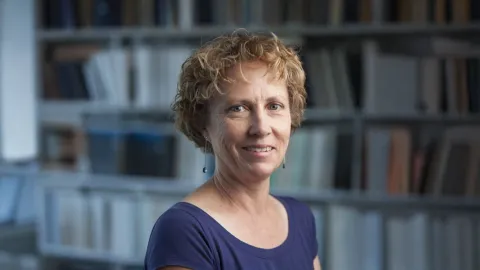 A color photo of woman with short curly hair. Books in background.