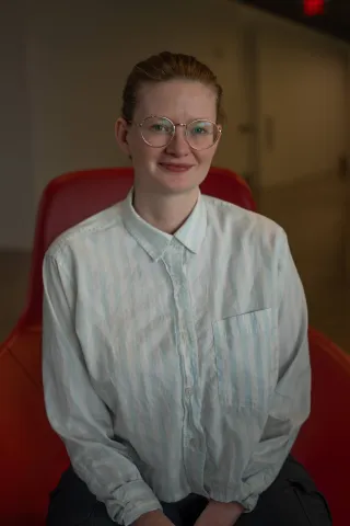A photo of Sarah Dean, a woman with glasses and blond hair pulled back and a white shirt, sitting in a red chair.