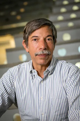A photo of Fred Schneider, a man with dark gray hair and a mustache, in a striped shirt in front of a staircase