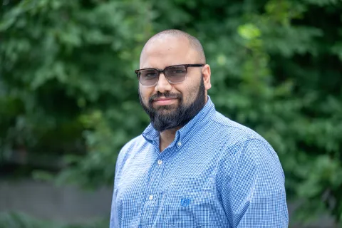 A photo of Sainyam Galhotra, a man with a shaved head, a dark beard, dark glasses and a blue shirt in front of a leafy background.
