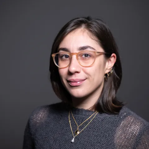 A headshot of Giulia Guidi, a woman with shoulder length brown hair, a gray sweater, brown glasses and gold necklaces.