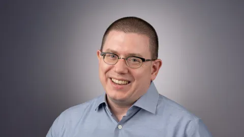 A color photo of Michael Clarkson, a smiling man with a buzzed head, glasses and a gray shirt in front of a gray background