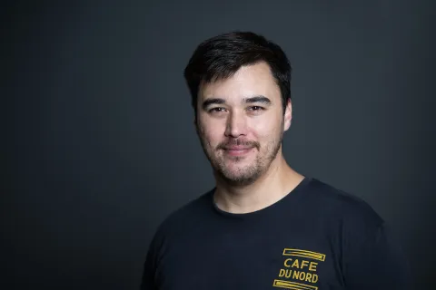 A photo of Michael Kim, a man with short black hair, a beard and mustache, in a black tshirt in front of a gray background