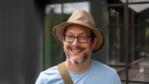 A photo of David Bindel, a man in glasses, a brown hat and blue tshirt