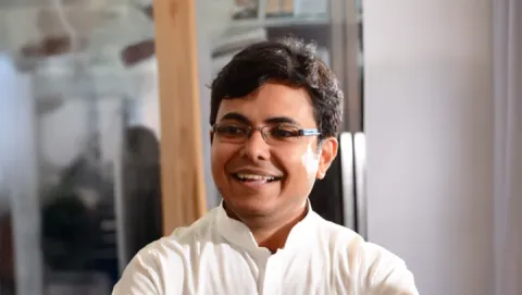 A photo of Karthik Sridharan, a smiling man with short dark hair, glasses and a white shirt