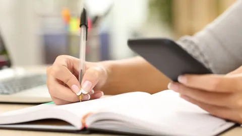 A color photo showing a person using a smart phone and writing down notes. Shutterstock