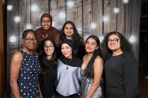 a group of female students poses together against a wooden-like backdrop with lights