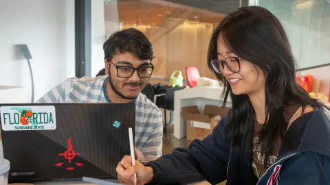 Two students sit around a laptop computer with a Flordia license plate bumper sticker. They wear glasses and smile, looking down at a tablet.
