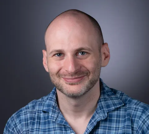A photo of Noah Stephens-Davidowitz, a smiling man with a shaved head, mustache and beard, and a blue plaid shirt in front of a gray background