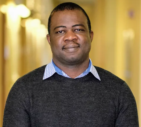 A photo of Owolabi Legunsen, a man with short black hair and mustache, in a black sweater in front of a gold background