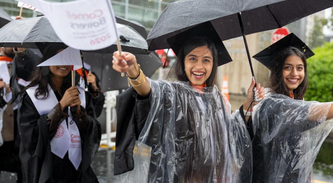 two female graduates walk under a umbrella and wave white pennants during processions