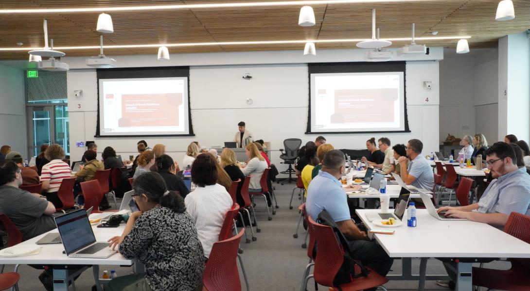 a classroom full of people sit at tables working on laptops