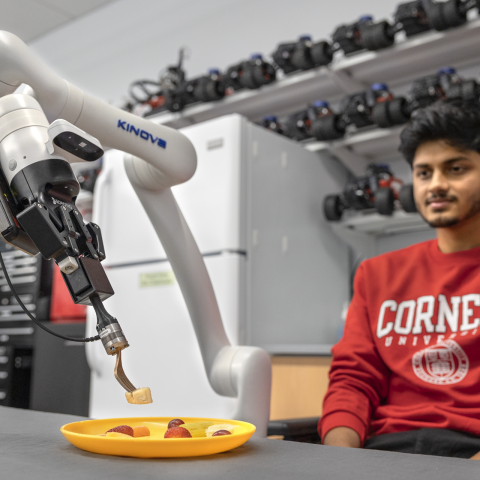 A color photo of a man sitting near a robotic arm using a fork to pick up fruit.