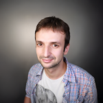 A headshot of a man with short brown hair, wearing a tshirt and button-down