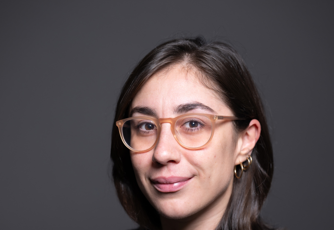 A headshot of Giulia Guidi, a woman with shoulder length brown hair, a gray sweater, brown glasses and gold necklaces.