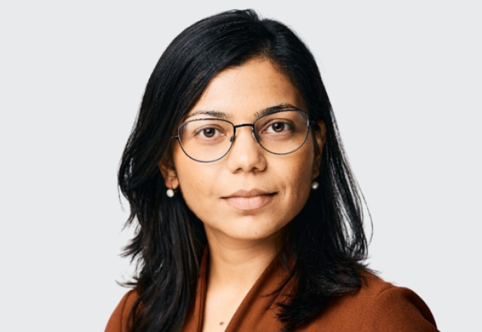 A photo of Rachee Singh, a woman with long dark hair and glasses in a brown sweater in front of a gray background
