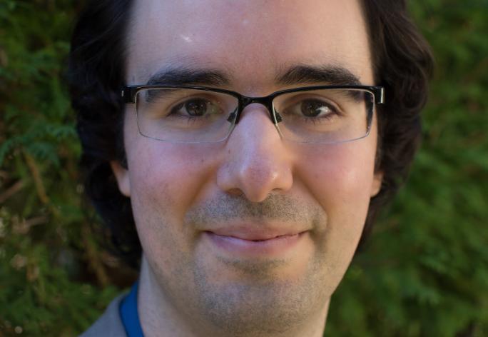 A photo of John Thickstun, a man with short black hair, glasses, and a blue shirt with a gray jacket in front of a leafy background