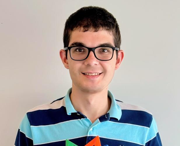 A photo of Matthew Eichhorn, a man with short dark brown hair, glasses, and a striped blue shirt, holding a colorful puzzle, in front of a gray background