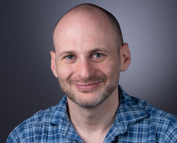 A photo of Noah Stephens-Davidowitz, a smiling man with a shaved head, mustache and beard, and a blue plaid shirt in front of a gray background