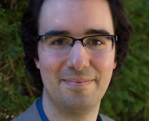 A photo of John Thickstun, a man with short black hair, glasses, and a blue shirt with a gray jacket in front of a leafy background