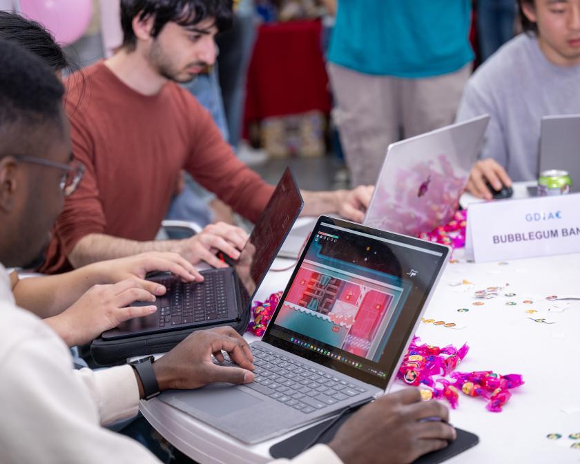 Four men sit at a round table playing videogames on laptops