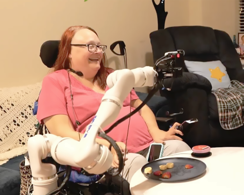 A color photo showing a woman using an assistive robotic device for eating.