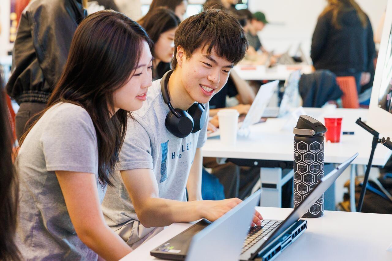 two students sit and work on laptops together